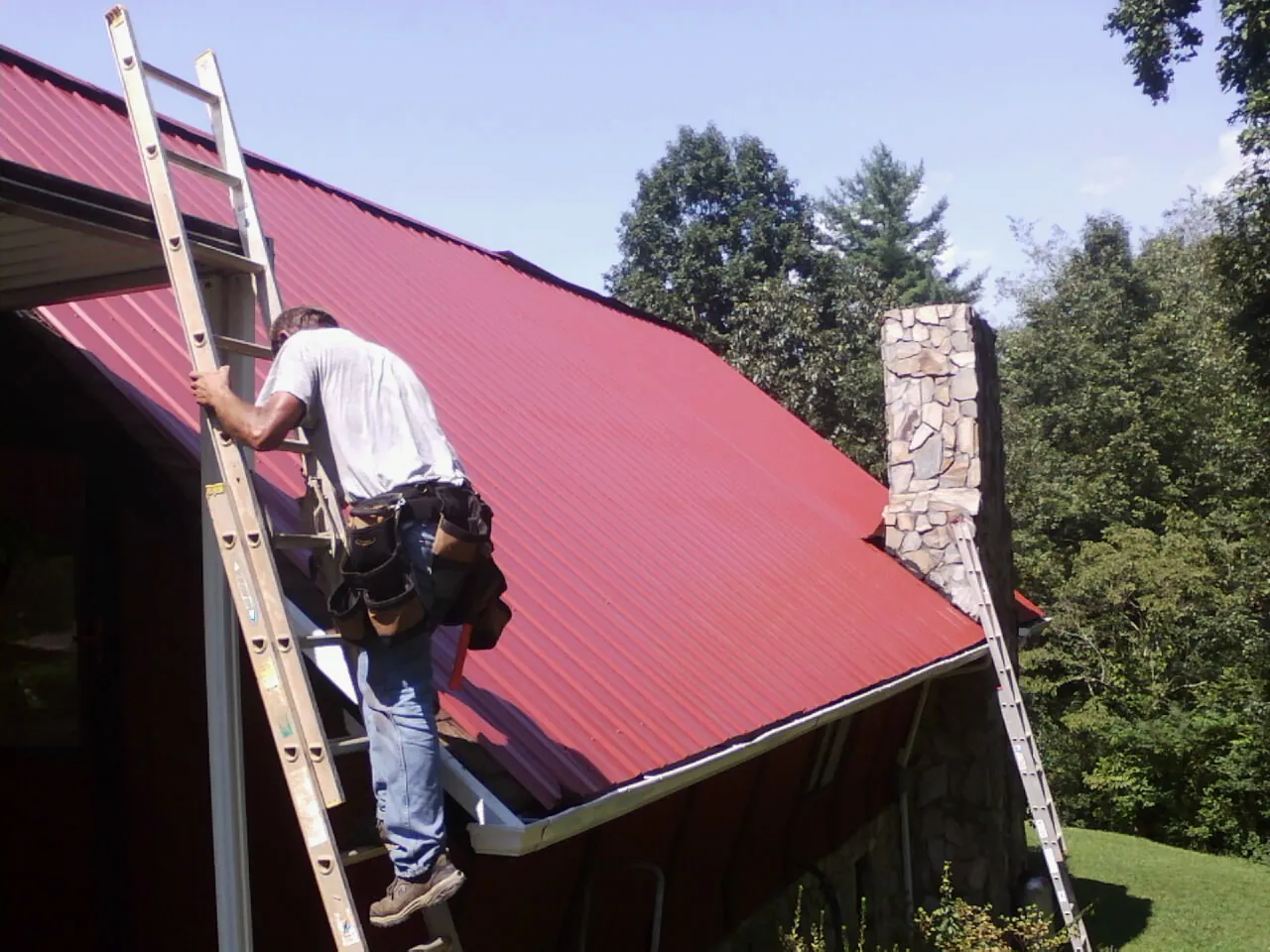 Roofer on a ladder beside a red metal roof and stone chimney — Soffit & Fascia Repair by Roofing Brothers in Sharpsville
